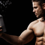 Model sports young man on dark background. Portrait of beautiful strong muscle guy with gray protein drink in shaker. Water splash.